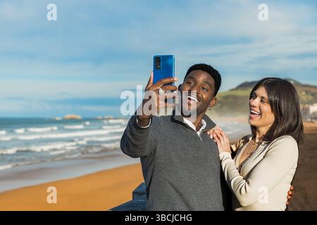 Ein lächelndes Paar genießt einen Moment zusammen und macht ein Selfie am Strand mit Meereswellen und Bergen im Hintergrund. Sie teilen Lachen und Glück Stockfoto
