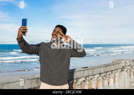 Fröhlicher Mann posiert mit einem Friedenszeichen, während er ein Selfie am Meer macht. Er lächelt hell vor der malerischen Kulisse der Wellen und des blauen Himmels, Capturi Stockfoto