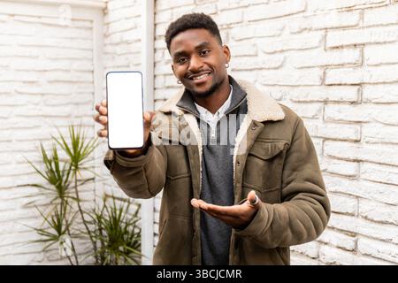 Ein junger Mann in einer warmen Jacke hält ein Smartphone mit einem leeren Bildschirm und bewegt sich mit einem freundlichen Lächeln darauf zu. Ideal für die Präsentation von Apps, Websites oder Stockfoto