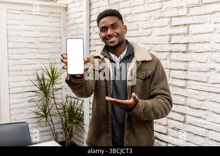 Fröhlicher junger Mann in einer warmen Jacke hält ein Smartphone mit einem leeren Bildschirm und zeigt mit einem verspielten Ausdruck darauf. Ideal für die Werbung für Apps, DIG Stockfoto