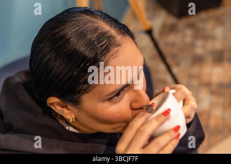 Eine Nahaufnahme eines Mädchens, das drinnen Kaffee schlürft. Sie hat dunkle Haare ordentlich gekämmt und trägt einen schwarzen Hoodie. Ihre goldenen Ohrringe und der rote nagellack Stockfoto