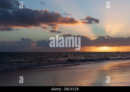 Eine beruhigende Ozeanszene bei Sonnenuntergang mit leuchtenden Sonnenstrahlen, die durch Wolken brechen. Ruhige Wellen erreichen sanft das Ufer und schaffen eine ruhige und dennoch dramatorige Atmosphäre Stockfoto