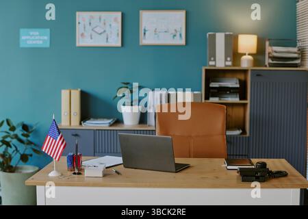 Visa-Center mit Holzschreibtisch mit organisiertem Zubehör, Laptop und amerikanischer Flagge. Die Wand ist mit farbenfrohen Kunstwerken und einer beleuchteten Lampe verziert, die Ihnen einen Vorgeschmack bietet Stockfoto