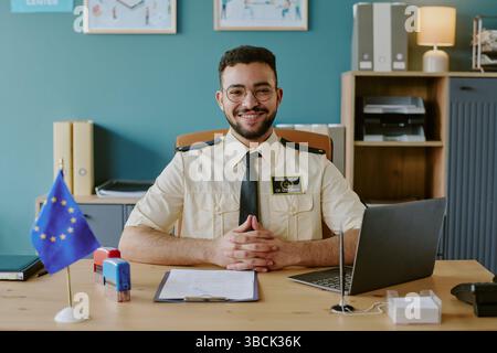 Porträt eines lächelnden Büroangestellten mit Ausweis und Brille, auf dem Schreibtisch mit Laptop und blauer EU-Flagge, Regalen mit Büchern und Unterlagen Stockfoto