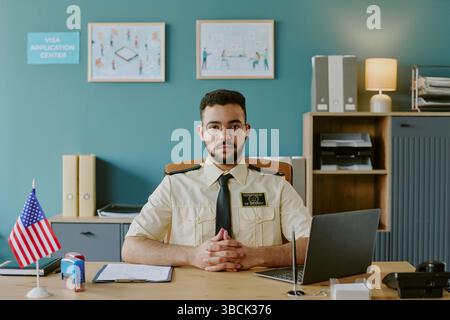 Porträt eines jungen Mannes, der am Schreibtisch im Visumantragszentrum sitzt, mit ernstem Ausdruck in die Kamera blickt, umgeben von Bürobedarf und USA fla Stockfoto