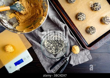 Kugeln mit Keksteig, gerollt in Sesam Seeds: Keksteig, weiße und schwarze Sesamsamen, Kekslöffel, Küchenwaage und Pergament gefütterte Blechpfanne Stockfoto