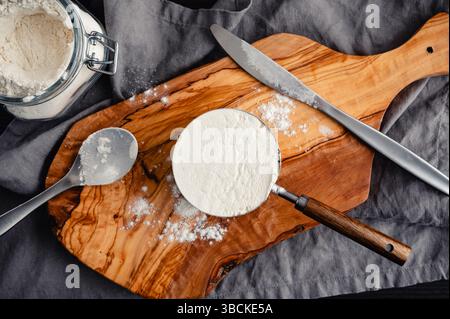 Messbecher mit Allzweckmehl: Direkt über einer eingeebneten Tasse Mehl mit Löffel, Messer, Schneidebrett und Mehl Stockfoto