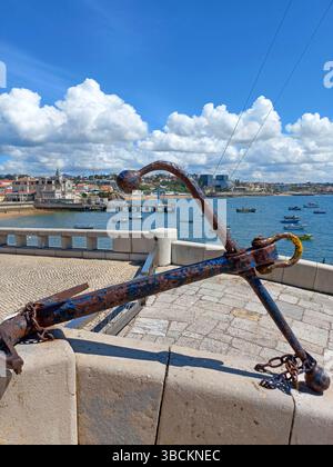 Alter maritimer Anker in Cascais Portugal auf einer Steinpromenade mit Blick auf die Hafenboote und die traditionelle Küstenstadt im Hintergrund Stockfoto