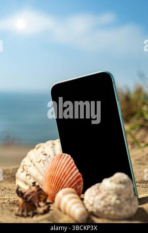 Vertikales Bild einer Nahaufnahme von Muscheln, die auf dem Sand einer kleinen Düne in der Nähe des Meeres angeordnet sind. Zwischen ihnen, ein Handy. Stockfoto