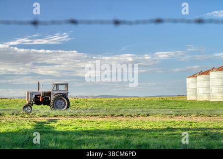 Der alte Traktor sitzt im Leerlauf auf einem grünen Feld neben Kornsilos mit einem Stacheldrahtzaun im Vordergrund Stockfoto