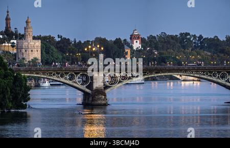 Die Dämmerung strahlt einen wunderschönen Glanz über die Triana-Brücke in Sevilla aus und hebt den Torre del Oro und die lebhafte Atmosphäre am Ufer hervor. Stockfoto
