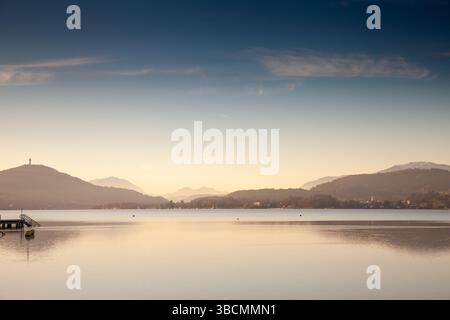 Die goldene Dämmerung siedelt sich über dem Worthersee bei Klagenfurt; Österreich. Ruhiges, spiegelähnliches Wasser spiegelt weit entfernte bewaldete Hügel und helle alpine Silhouetten wider Stockfoto