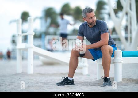 Sport-Fitness-Mann, der draußen auf einer Bank ruht. Reifes Fitnesssportmodell, das nach dem Training auf einer Bank sitzt. Ein Sportmann macht nach dem Fitnesstraining eine Pause Stockfoto