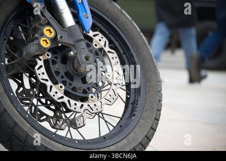 Detaillierte Ansicht eines Motorrads mit sichtbarer Scheibenbremse und Reifen, umgeben von einem verschwommenen urbanen Hintergrund mit Fußgängern und Gehsteig Stockfoto