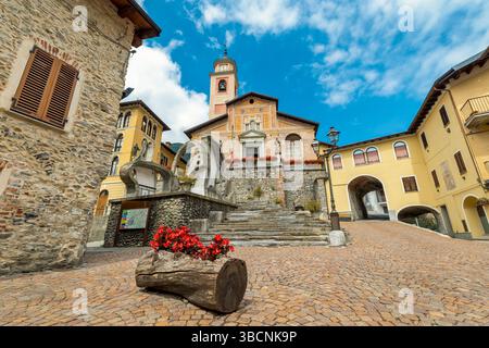 Historische Kirche und umliegende Gebäude auf einem kopfsteingepflasterten Stadtplatz unter einem teilweise bewölkten blauen Himmel in Entraque, Italien. Stockfoto