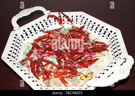 Eine lebendige Kollektion getrockneter roter Chilis in einem wunderschön gestalteten weißen Korb mit kunstvollen Griffen. Das Tablett hat farbenfrohe Blumenmotive. Stockfoto