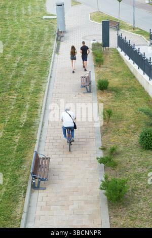 Ein älterer Mann radelt mit dem Fahrrad, während ein junges Paar einen gepflasterten Stadtparkweg entlang spaziert, der von Bänken und Gras bei hellem Tageslicht umgeben ist. Stockfoto