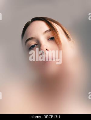 Konzeptionelle Kunstwerke. Porträt einer rothaarigen Frau mit durchdringenden blauen Augen, die durch den verschwommenen Filter nach unten blickt, Isolation vermittelt und Stockfoto