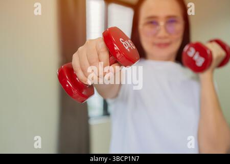 Asiatische Frau mittleren Alters lächelt selbstbewusstes Training mit Kurzhanteln zu Hause. Fit und gesund halten während der Quarantäne. Stockfoto