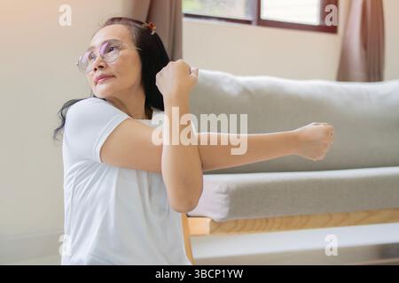 Glückliche und gesunde ältere asiatische Frau, die zu Hause eine Schulterbewegungsübung macht und mit einem Querarm-Stretch arbeitet. Stockfoto
