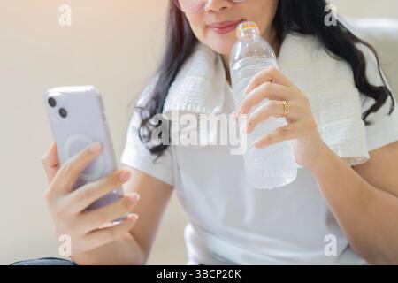 Müde Seniorin mit Wasserflasche und Telefon in der Hand, während sie sich nach dem Heimsport-Training entspannt. Relax-Konzept. Stockfoto