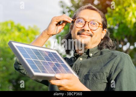 Überraschter erwachsener asiatischer Mann mit tragbarem Solarmodul, der seine eigene Energie auf grünem natürlichem Hintergrund auflädt. Lustiges Gesicht. Selektiver Fokus. Stockfoto