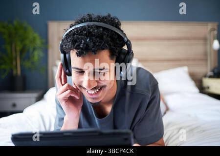 Glücklicher junger Mann, der Musik über Kopfhörer genießt, während er sich auf einem Bett zu Hause entspannt Stockfoto