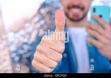 Ein lächelnder Mann zeigt Daumen nach oben mit Smartphone in der Hand, Nahaufnahme-Gestenfokus Stockfoto