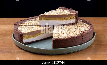 Dekadenter, mehrschichtiger Kuchen mit Kakaokruste, Vanille- und Schokoladenmousse, knuspriger Erdnussschicht und Nussgarnitur. Schnitt auf Platte angehoben, Studioschoss gegen Stockfoto