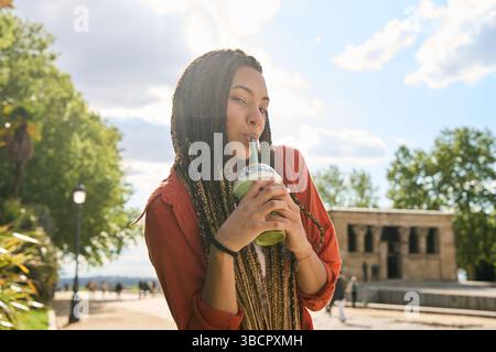 Junge Frau mit langen Zöpfen, die an einem sonnigen Tag im Park ein kaltes Getränk genießt, mit dem Tempel von debod im Hintergrund Stockfoto