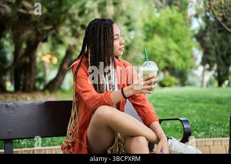 Junge Frau mit langen Zöpfen, die auf einer Bank in einem Park sitzt, einen erfrischenden Matcha Latte hält und das Sommerwetter genießt Stockfoto
