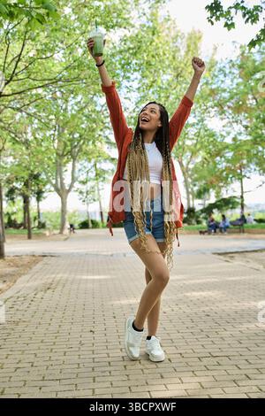 Unbeschwerte junge Frau mit langen Zöpfen, die den Sommer in einem Park fröhlich feiert, einen leuchtenden grünen Smoothie hält und den sonnigen Tag begrüßt Stockfoto