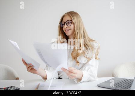 Junge Frau, die Dokumente an einem modernen Schreibtisch an einem hellen Tag in einem minimalistischen Arbeitsbereich liest Stockfoto