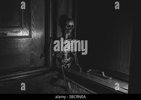 Großes Skelett in der Ecke neben der Tür auf dem Boden im Dunkeln, gruseliges Skelett, Schwarzweißfoto Stockfoto