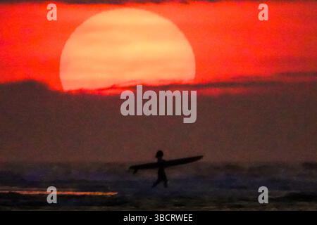 Folly Beach, Usa. Mai 2025. Eine Surferin sieht den Sonnenaufgang, als sie am 21. Mai 2025 in Folly Beach, South Carolina, zum Surf am Strand, bekannt als The Edge of America, spaziert. Quelle: Richard Ellis/Richard Ellis/Alamy Live News Stockfoto