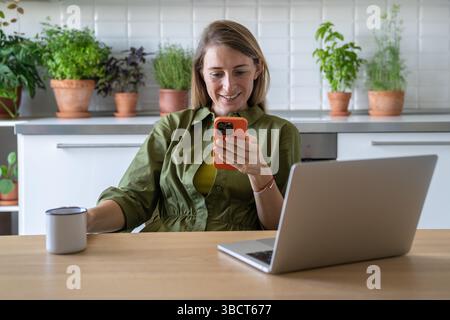 Fröhliche Frau, die in der Küche Pause macht, auf Smartphone schaut, im Internet läuft und Tasse Tee hält Stockfoto