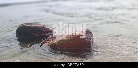Sanfte Wellen, die über zwei rötliche Felsen an einem Sandstrand schwimmen Stockfoto