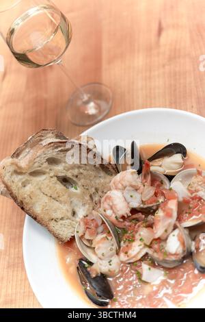 Cioppino Seafood Suppe mit Brot // Eine großzügige Portion Cioppino, die klassische italienisch-amerikanische Meeresfrüchtesuppe, wird in einer weißen Schüssel mit mehreren Scheiben frisch geröstetem Brot serviert. Die hellrote Tomatenbrühe enthält eine große Vielfalt an Meeresfrüchten, darunter Garnelen, Weißfisch, Muscheln mit Schwarzschalen und Muscheln. Dieser herzhafte Fischeintopf zeigt die natürlichen Farben der Meeresfrüchte und die rustikale Tomatenbrühe, wobei das geröstete Brot zum Eintauchen in die aromatische Flüssigkeit positioniert wird. Stockfoto
