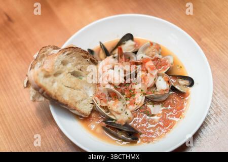 Cioppino Seafood Suppe mit Brot // Eine großzügige Portion Cioppino, die klassische italienisch-amerikanische Meeresfrüchtesuppe, wird in einer weißen Schüssel mit mehreren Scheiben frisch geröstetem Brot serviert. Die hellrote Tomatenbrühe enthält eine große Vielfalt an Meeresfrüchten, darunter Garnelen, Weißfisch, Muscheln mit Schwarzschalen und Muscheln. Dieser herzhafte Fischeintopf zeigt die natürlichen Farben der Meeresfrüchte und die rustikale Tomatenbrühe, wobei das geröstete Brot zum Eintauchen in die aromatische Flüssigkeit positioniert wird. Stockfoto