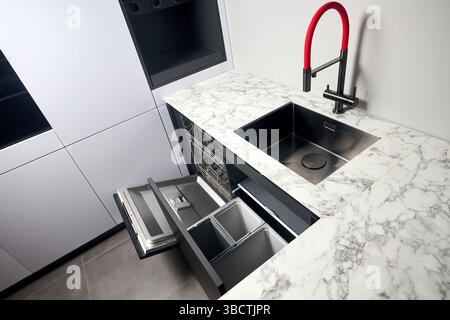 Küche mit eingebautem, geöffnetem Geschirrspüler und herausziehbarer Schublade für die getrennte Abfallsammlung. Wasserhahn Waschbecken mit kompaktem Hochdruck Stockfoto