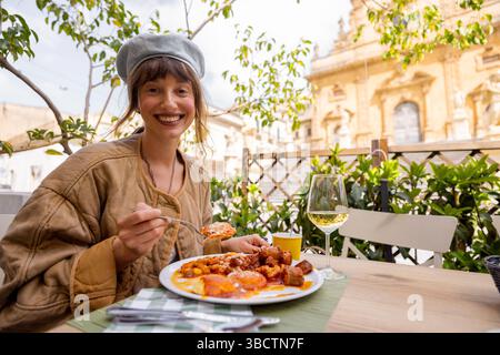 Die lächelnde Frau genießt ein herzhaftes sizilianisches Mittagessen mit Gnocchi, Fleischbällchen und Tomatensoße, gepaart mit Weißwein, in einem Restaurant im Freien vor der Petersdom in Modica Stockfoto