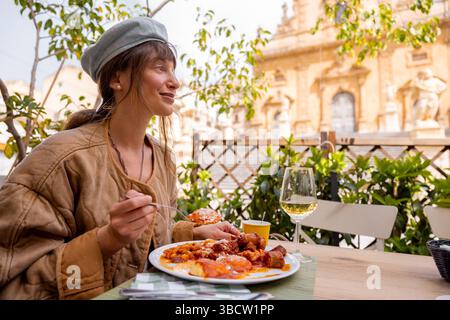 Die lächelnde Frau genießt ein herzhaftes sizilianisches Mittagessen mit Gnocchi, Fleischbällchen und Tomatensoße, gepaart mit Weißwein, in einem Restaurant im Freien vor der Petersdom in Modica Stockfoto