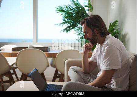 Ein Mann, der auf einer Couch in einem modern eingerichteten Wohnbereich sitzt und sorgfältig an einem Laptop arbeitet, verkörpert Konzentration und Hingabe in einer ferngesteuerten Arbeitsumgebung Stockfoto