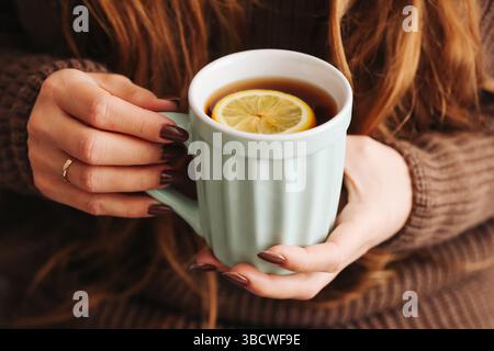 Frau mit heißem Tee. Mintgrüner Becher mit Zitronenscheibe. Kuscheliges braunes Pullover-Outfit. Entspannender Moment mit warmem Getränk. Winterkomfort am Morgen. Stockfoto