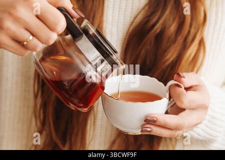 Tee aus dem Topf gießen. Frau mit elegantem Becher. Morgens wird ein warmes Getränk serviert. Gemütliche Teezeit zu Hause. Aromatischer schwarzer Tee. Tragekomfort und Wärme im Winter. Stockfoto