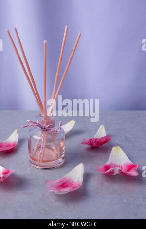 Ein zarter Aroma-Diffusor sitzt umgeben von weichen rosa Blüten auf einer ruhigen, glatten Oberfläche mit Lavendelhintergrund. Stockfoto