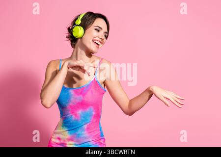 Die charmante junge Frau tanzt fröhlich in einem farbenfrohen Sommerkleid vor rosa Hintergrund und verkörpert lebendige Mode Stockfoto