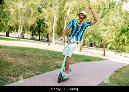Junger Mann in gestreiftem Hemd, der einen sonnigen Tag mit einem Roller im Park genießt Stockfoto