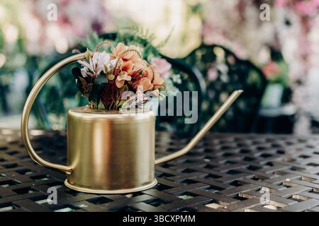 Goldene Gießkanne mit künstlichen Blumen in Pastellfarben auf dem Gartentisch, die ein gemütliches, florales Stillleben mit zarten Farben und sanftem Fokus schaffen Stockfoto