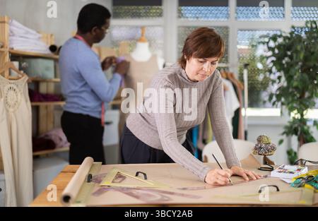 Die Schneiderin zeichnet in der Nähwerkstatt anhand von Mustern eine Skizze der zukünftigen Kleidung auf Papier Stockfoto